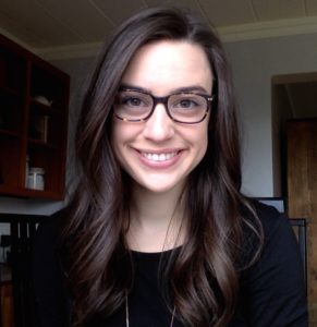Tori has long brown hair, is wearing tortoise-shell glasses, a black shirt, and is smiling.