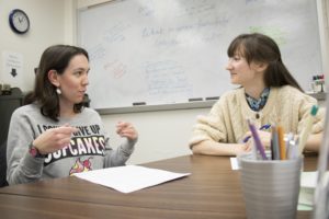 A K-State student (left) discusses her writing, using hand gestures, while a tutor (right) listens and makes eye contact.