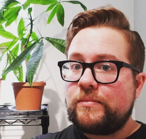 Neil looks forward in black frame glasses with a small money plant on a shelf to his left.