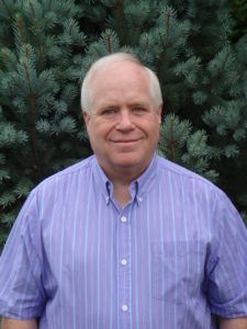 Brad Hughes wears a purple hue button up shirt and smiles in front of pine trees.