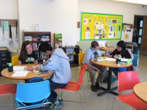 A pair of students and tutors gather at round tables peering down at pieces of writing.