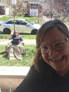 Close-up of a smiling Stephanie outside, with a colleague seated on her lawn in the distance, waving and smiling.