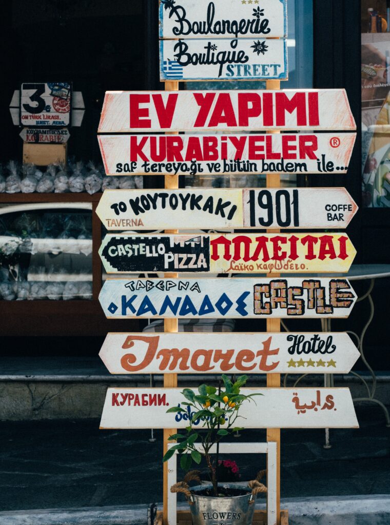 This photo by Soner Eker on Unsplash shows a series of signs directing the viewer to different establishments located in Kavala, Greece. A plant sits in a silver bucket marked "Flowers" below.