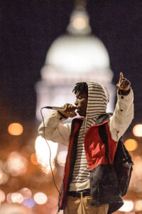 A Black student speaks to a crowd while holding a microphone in front of the Wisconsin capital building.