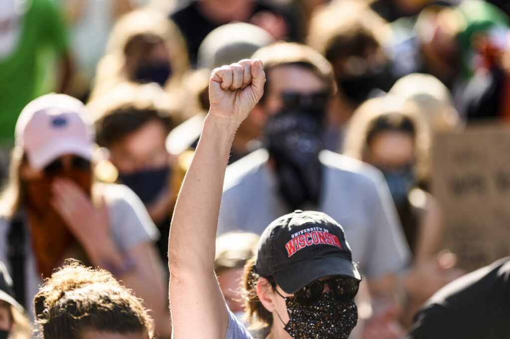 A white woman wearing a University of Wisconsin hat, sunglasses, and a mask, raises her fist in the air at a Black Lives Matter solidarity march