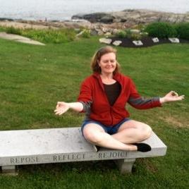 A woman meditates while sitting cross-legged on a bench near the ocean.