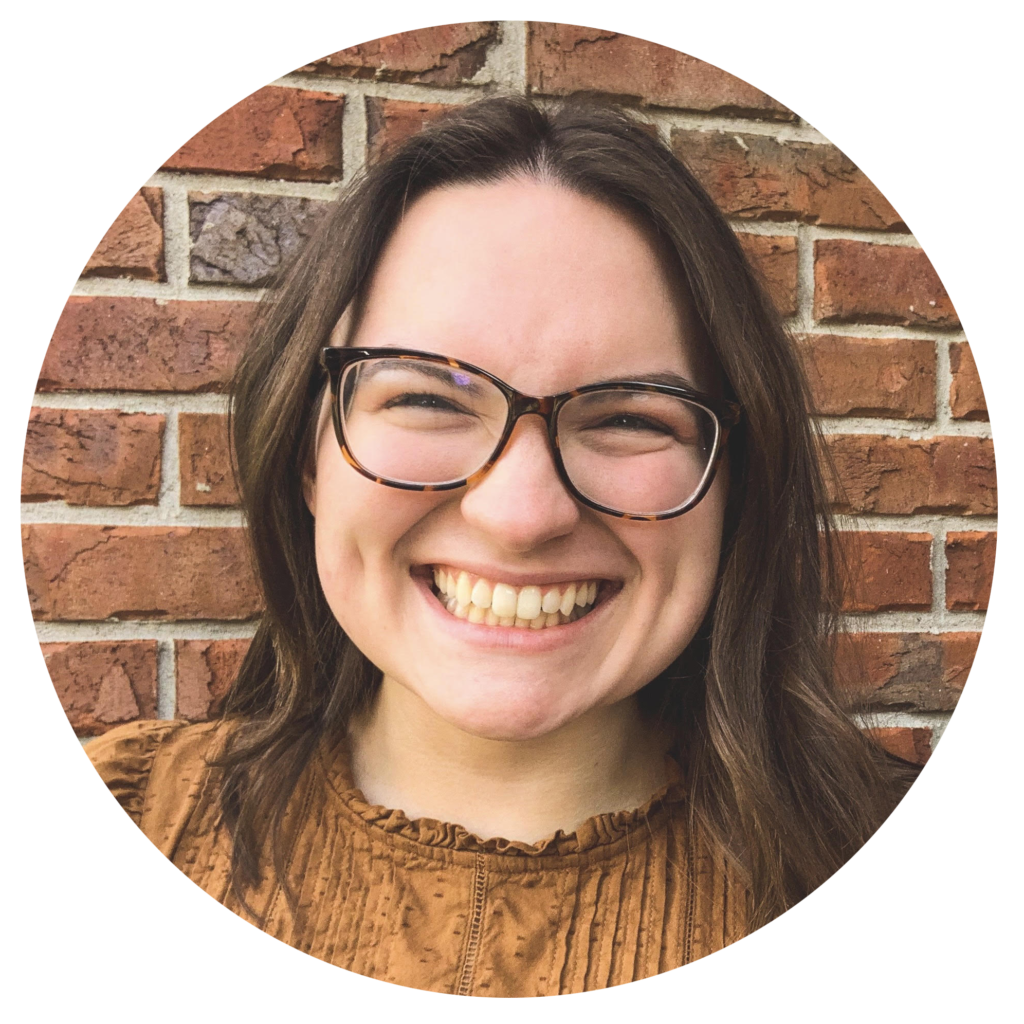 Author Caroline Shutt, wearing tortoiseshell-framed glasses and standing in front of a brick wall, smiles at the camera.