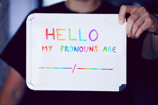 A white board with the text "HELLO MY PRONOUNS ARE __/___" in rainbow colors held in front of a person who is off camera