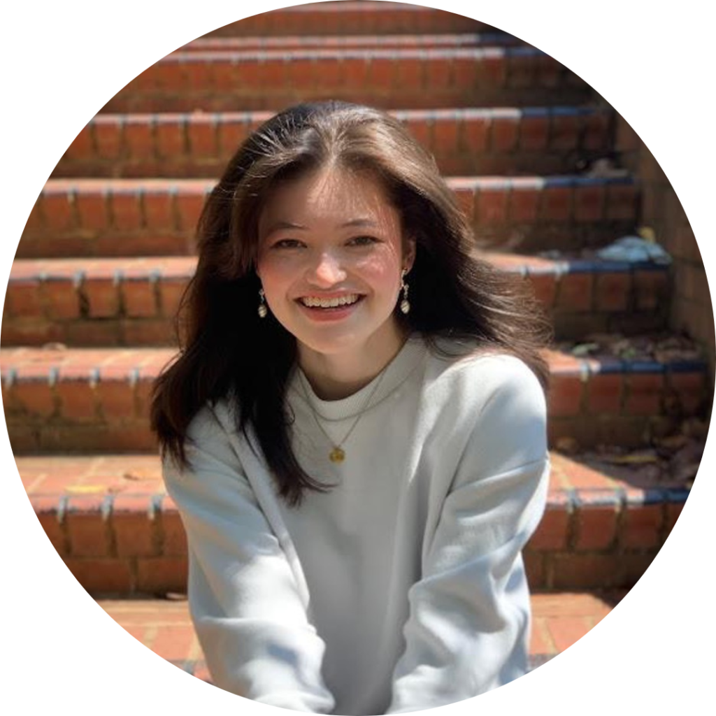 Maya is sitting on a brick staircase. She is wearing blue jeans, a gray long sleeve shirt, gold earrings, and a gold necklace.