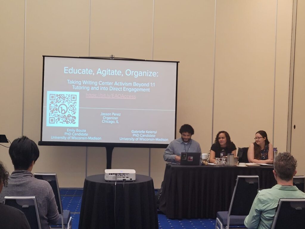 Emily Bouza, Gabrielle Kelenyi, and Jasson Perez sitting in front a conference room. There's a display screen in the background that shows a title slide for the workshop "Educate, Agitate, Organize: Taking Writing Center Activism Beyond 1:1 Tutoring and into Direct Engagement."