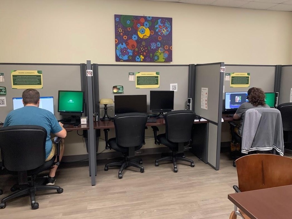 Three open-backed cubicles against the wall, side by side. Each cubicle has a desk, two computer monitors, and two chairs. The middle cubicle is empty, while the two that flank it are occupied by tutors. Above the middle cubicle is a large painting of flowers. Each cubicle has informational flyers stapled to its walls.