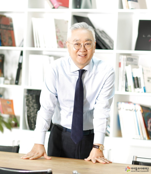 Michael Lee stands behind a conference table with a wall of bookshelves behind, leaning forward and smiling at the camera.