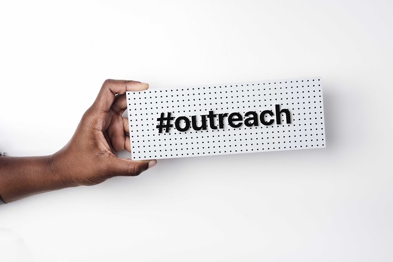 A hand holding a white sign with a hashtag and the word outreach