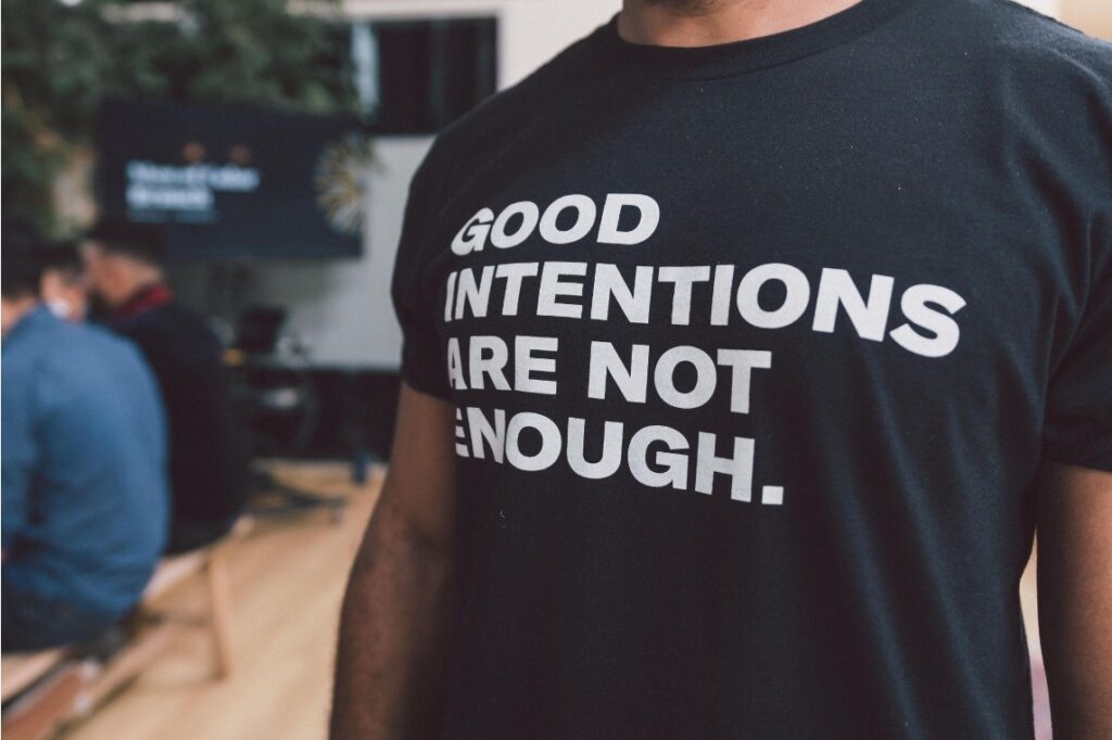 A Black man wearing a shirt that reads “Good Intentions are not enough.”