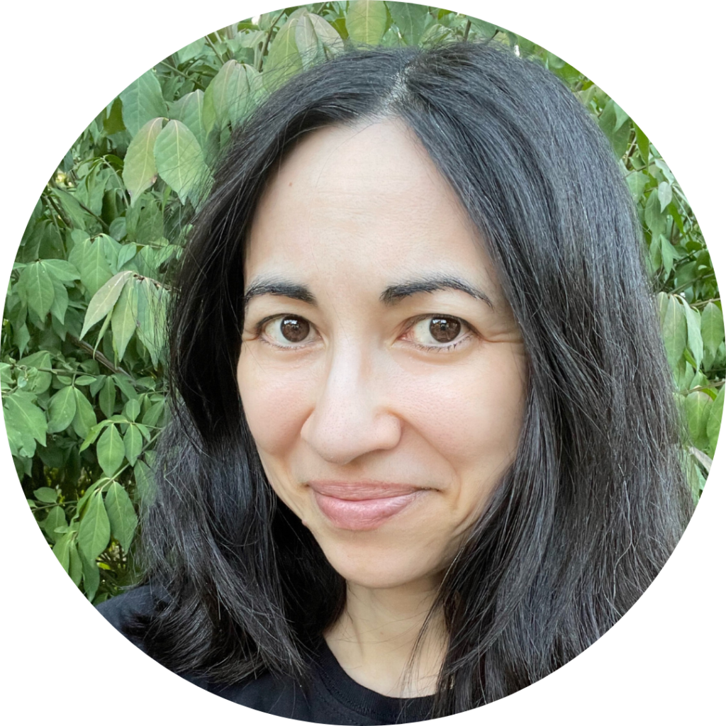 Rachel Azima with shoulder length dark hair, brown eyes, and a black t-shirt, smilling slightly at the camera, with green leaves in the background.
