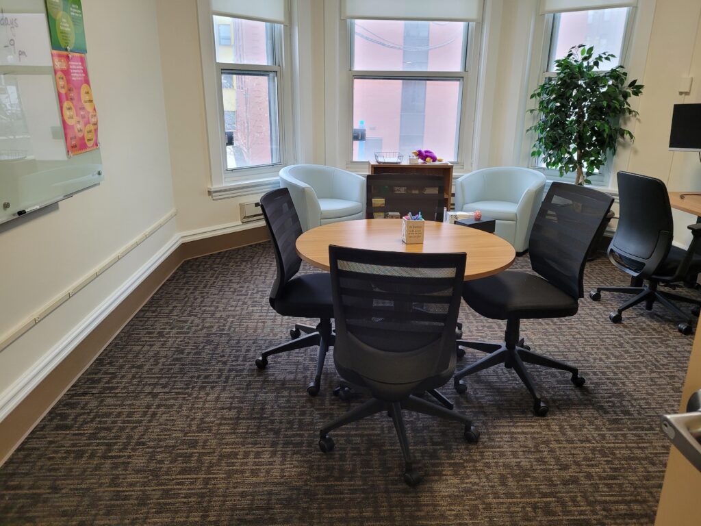 a bright, carpeted room with 3 large windows, two comfortable chairs, a small round table with four office chairs around it, a white board on freshly painted walls, and a large plant in the corner