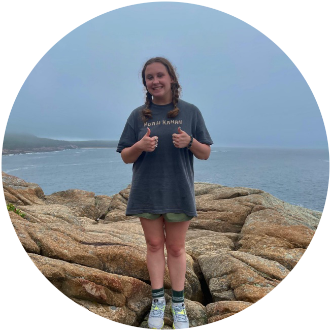 Grace Apostol gives two thumbs up as she stands on a rocky coastline.