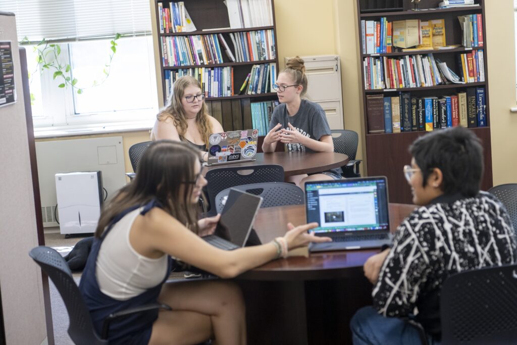 Two pairs of students are in writing center sessions, talking and gesturing over shared laptops. 