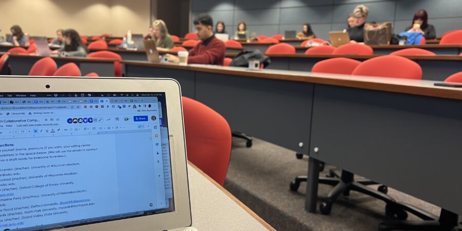 A computer with writing on it in front of a classroom filled with writing center professionals writing