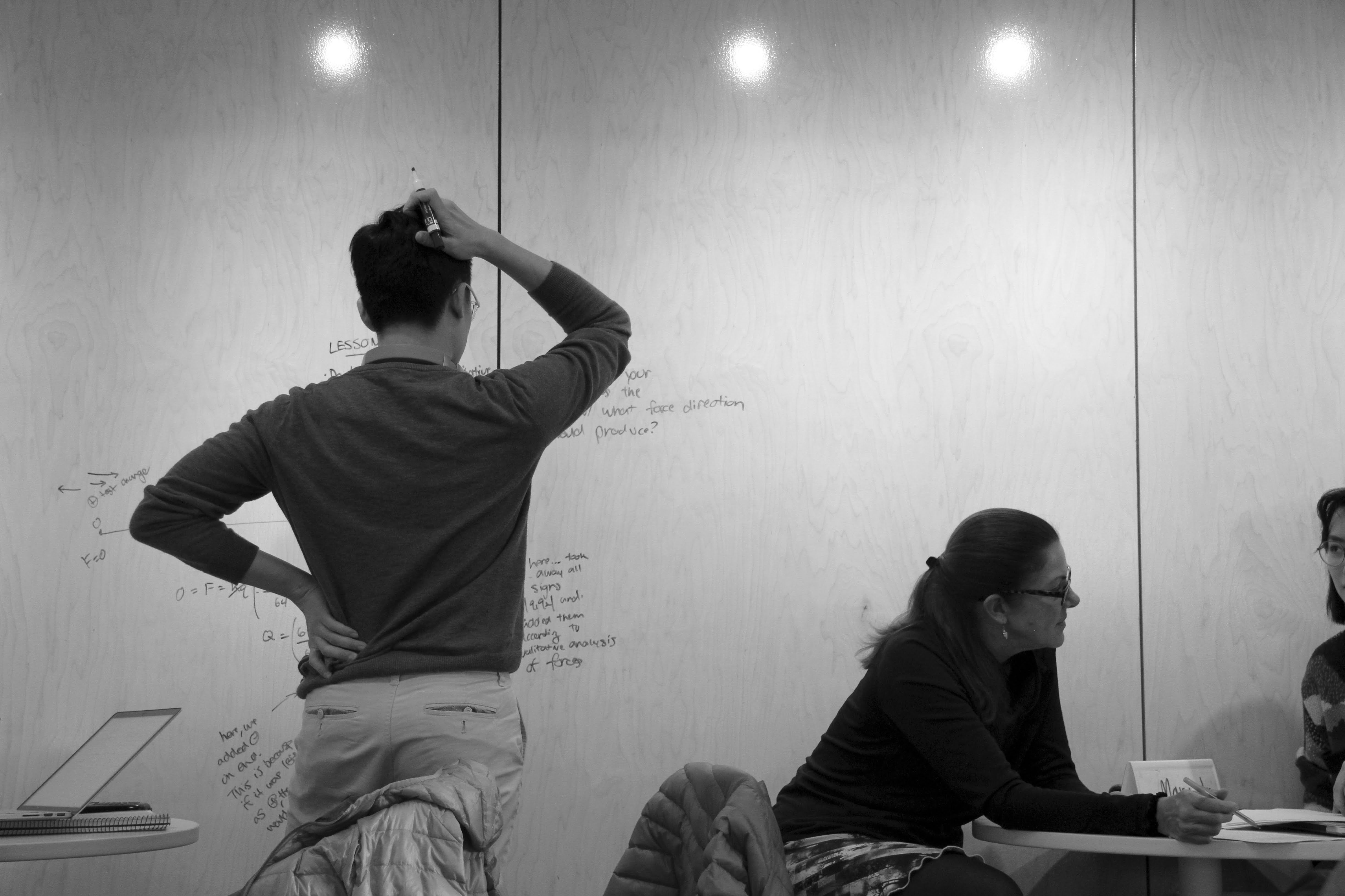 One student with his back to the camera, one hand on his left hip, the other scratching his head as he stares at a writable wall filled with notes. To the right sit two other people at a table engaged in conversation, one of them gesturing at a paper between them. 