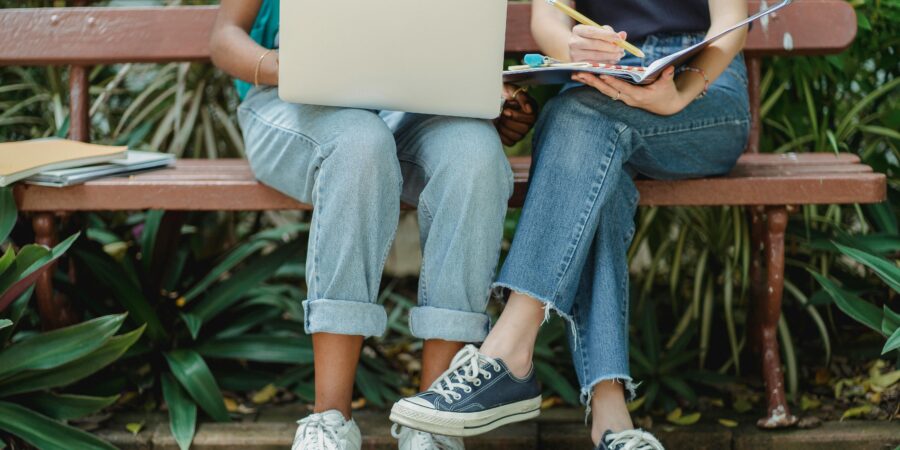 Two people sitting on a bench with a laptop and notebook on their laps.