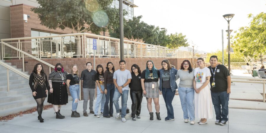 Thirteen multiracial people stand in a line in front of a campus building