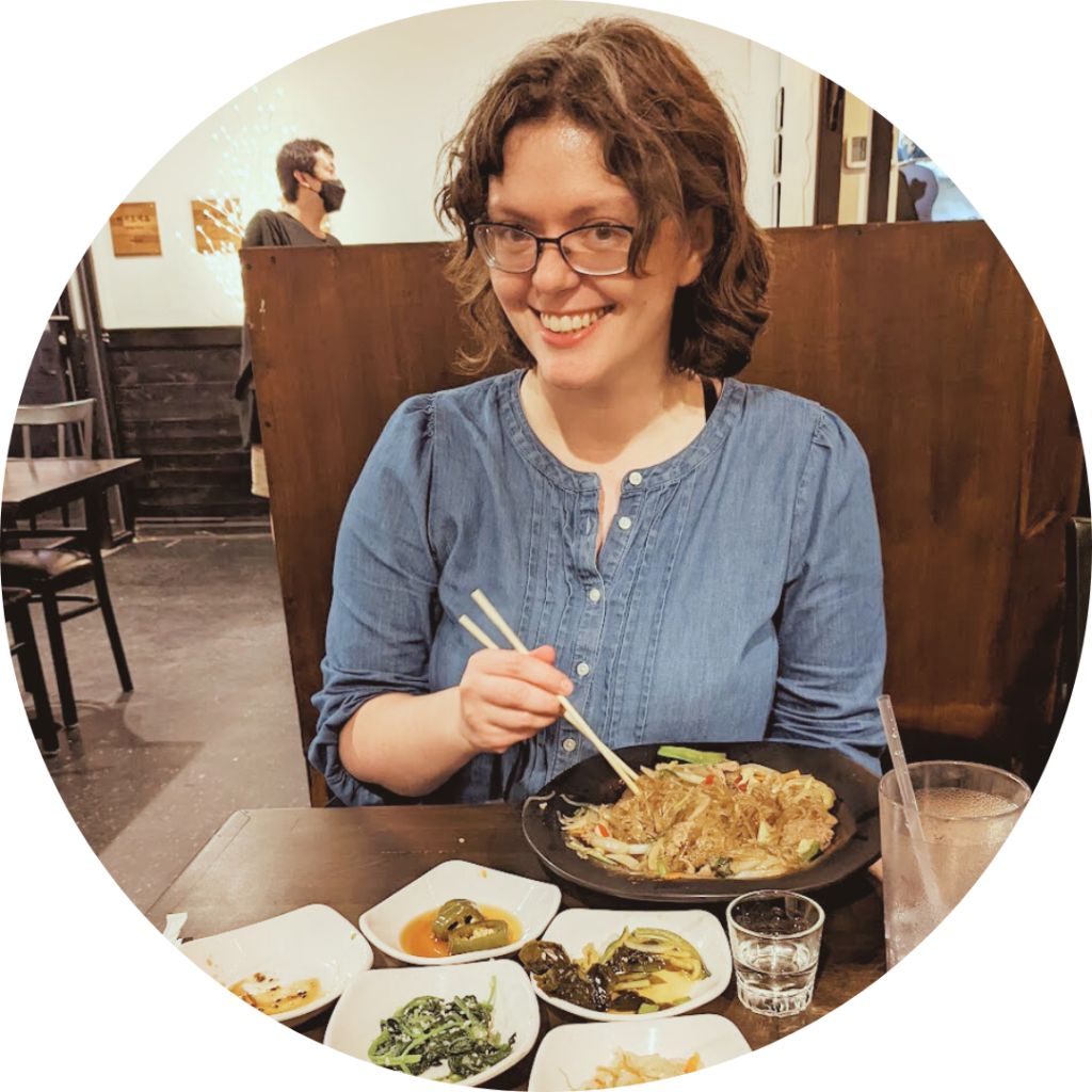 A white woman with brown hair and glasses sits in a restaurant eating bachan and smiling.