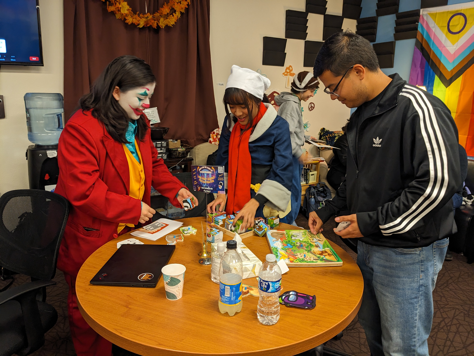 Three colleagues stand at a table playing with puzzles. One wears a joker costume.