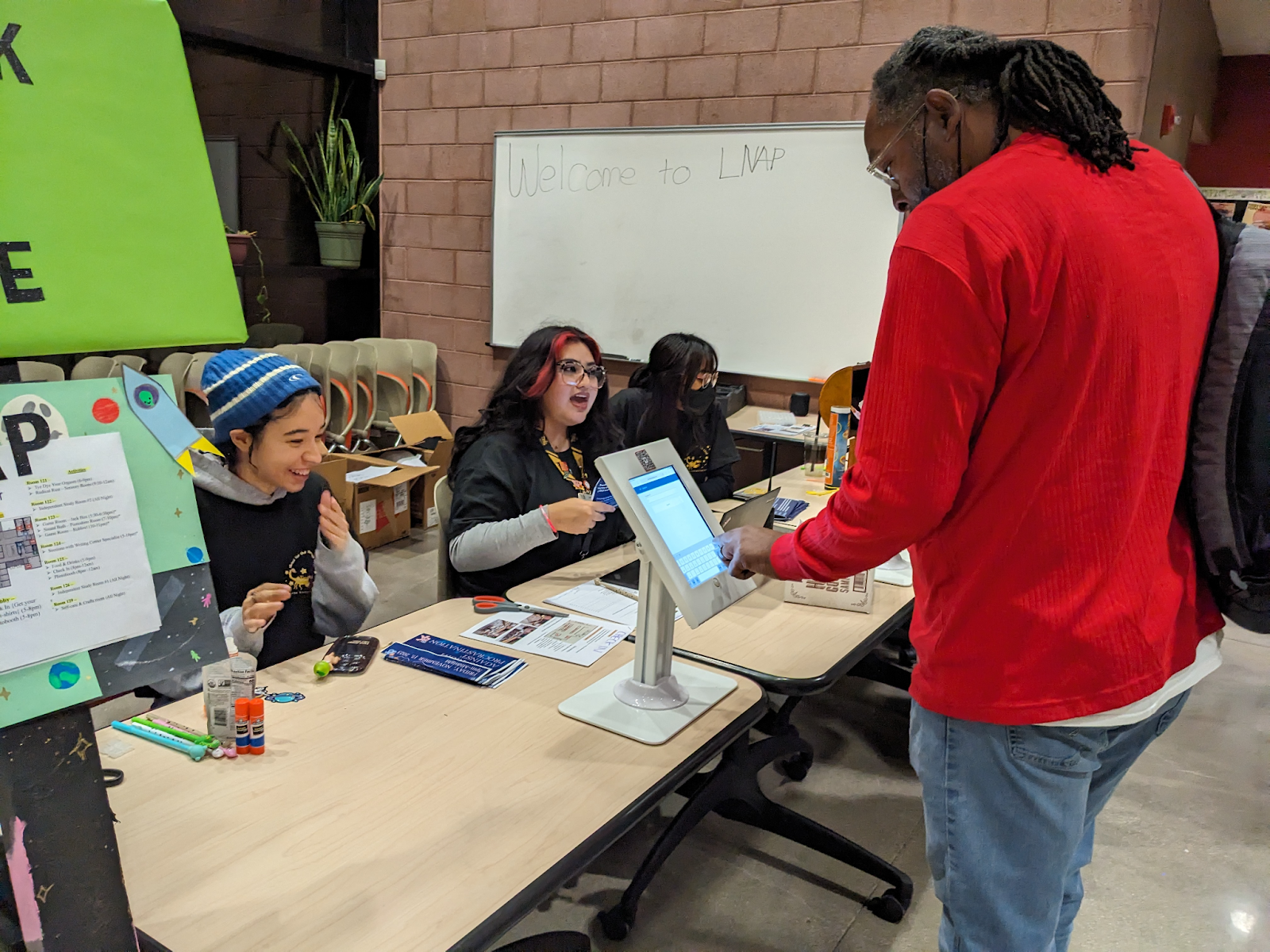 Three student-colleagues sit behind a table as one standing person checks-in on an ipad.