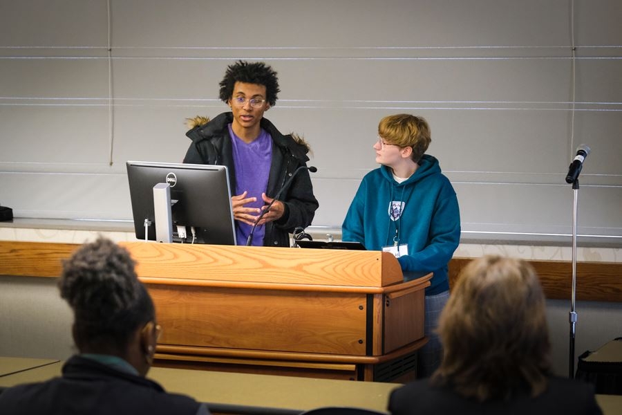 A Black woman on the left and a white man on the right stand together behind a podium; the woman is speaking to an audience visible by the backs of their heads.