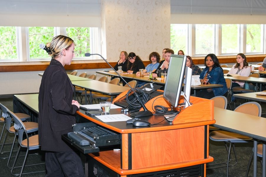 A white woman stands behind a podium and speaks into a microphone before an audience of several people.