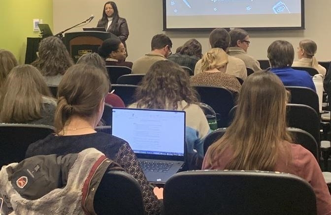 A South Asian woman speaks from behind a podium to a large audience visible from behind. One audience member has a page of notes open on their laptop.