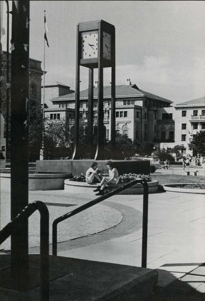 black and white photo of clock tower and two people visible sitting in the State Street Pedestrian Mall near Memorial Library on the UW-Madison campus