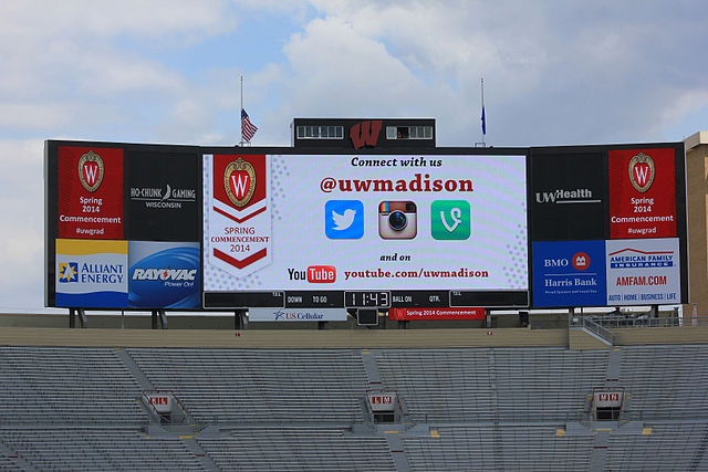 photograph of billboard from UW-Madison's 2014 Commencement, with a billboard advertising social media information (Connect with us @uwmadison) appearing over empty stadium seats