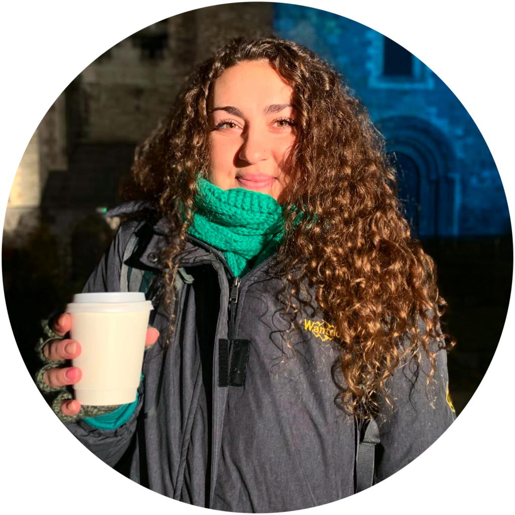 Meg, a young, white woman with curly brown hair, smiling at the camera, bundled in a green scarf and gray coat.