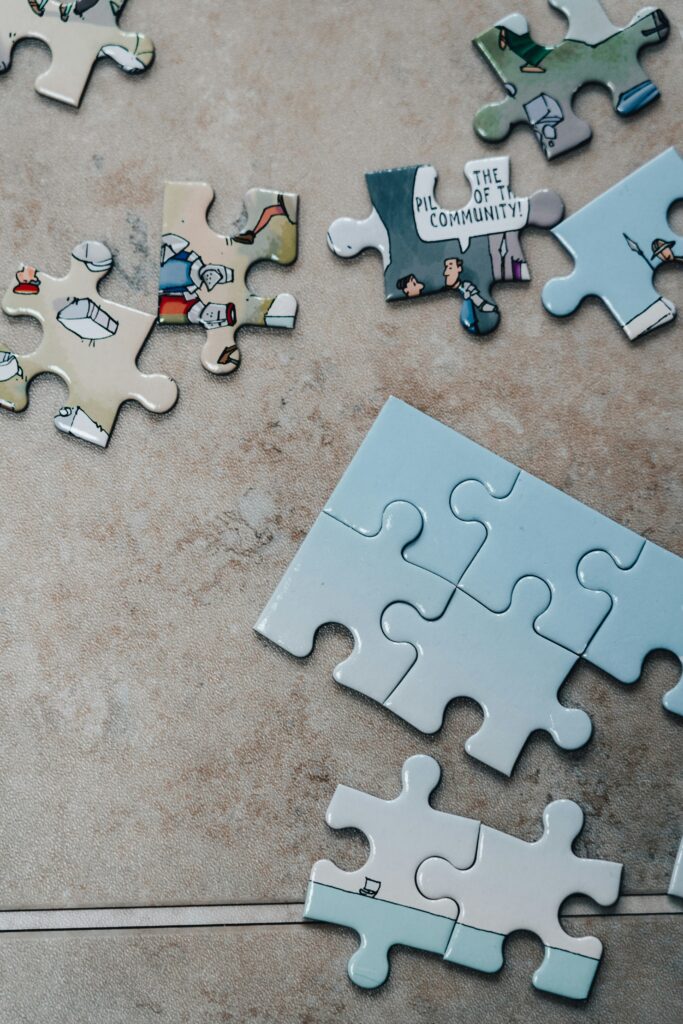 This photo pictures 13 puzzle pieces half of which are a faded blue color and the other half of which have cartoon looking designs on them with one saying “The Pillar of The Community.” The puzzle pieces are laid out with six pieces scattered at the top of the frame, five pieces connected in the middle of the frame, and two pieces connected at the bottom of the frame.