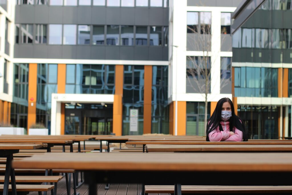 A young woman wearing mask sits alone on an empty bench outside, in the backyard of the office building.