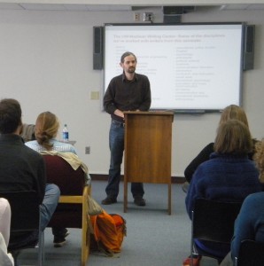 A photo of Dr. Paul Silvia standing at a podium in front of a projection screen, presenting to Writing Center tutors. Several people sit in chairs in the audience with their backs to the camera.