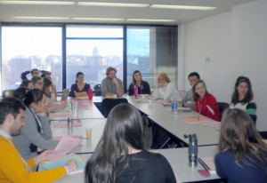 A series of tables aligned into a rectangle with teaching assistants sitting around them. There's papers, books, and pens on the table.