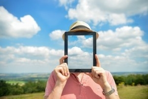 Person in a sunny field holding an ipad that reflects the clouds behind them.